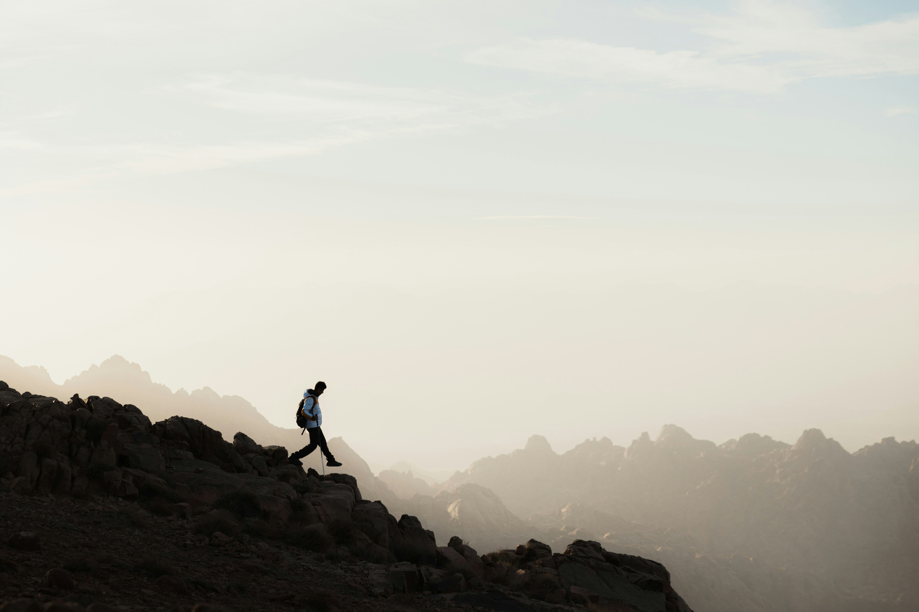 Auto-generated description: A person with a backpack is walking along a rocky ridge with a backdrop of misty mountains under a pale sky.